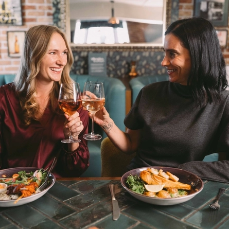 Two diners raising glasses of wine at a pub table, with a grilled skewer and rice dish and a fish dish in front of them.
