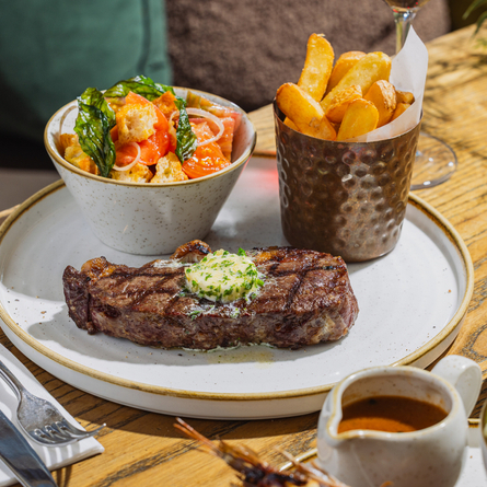 An advertising image showing 2 plated steak dishes sat on a table within the interior restaurant seating area at The Four Oaks.