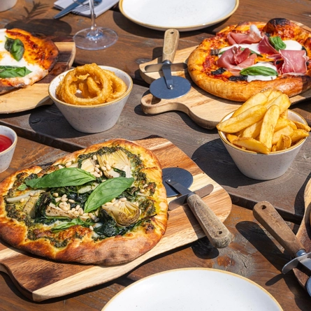 Various Pizzas on boards with bowls of Fries and Onion Rings at a Crafted venue.