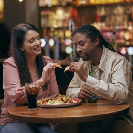 A man and woman sitting at a table with a bottle of Heineken and a glass of Coca Cola, sharing a plate of nachos.