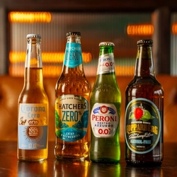 Four alcohol-free drinks are displayed on a wooden table in a pub setting: Corona Cero lager, Thatchers Zero cider, Peroni Nastro Azzurro 0.0%, and Kopparberg Strawberry & Lime Alcohol-Free cider. The bottles are arranged side by side against a softly blurred background with warm amber lighting.