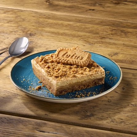 A close up view of a Biscoff Slice topped with a Lotus Biscoff biscuit, served on a plate which sits on a wooden table along with a spoon.