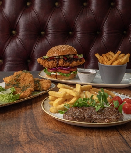 A plate of steak and chips, a plate of chicken kiev and fries, and a plate of Korean chicken burger and fries, sitting on a wooden table.