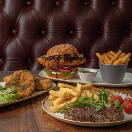 A plate of steak and chips, a plate of chicken kiev and fries, and a plate of Korean chicken burger and fries, sitting on a wooden table.