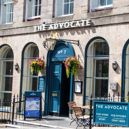 The exterior facade, signage and seating area of The Advocate in Edinburgh, with hanging flower baskets on either side of the entrance.