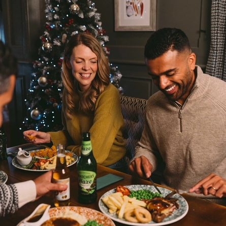 A group of people seated at a festive decorated table near a Christmas three. There are plates of food and drinks in front of them.