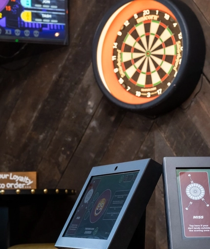 The darts area inside the Clocktower in Milton Keynes, with a dartboard, a TV on the wall, and a close up view of the electronic scoring system sitting on a table.