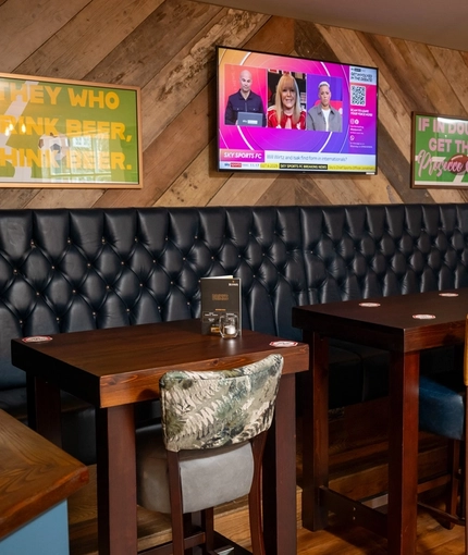 An interior seating area at The Kittoch in East Kilbride, with a long, upholstered booth seat, a TV on the wall above it, and wooden tables with upholstered bar stools.