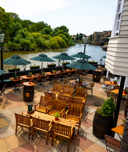 An aerial view of the paved, exterior beer garden seating area at The London Apprentice which overlooks the River Thames, with wooden tables and chairs, shade umbrellas, string lights, and planter boxes filled with flowers.
