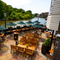 An aerial view of the paved, exterior beer garden seating area at The London Apprentice which overlooks the River Thames, with wooden tables and chairs, shade umbrellas, string lights, and planter boxes filled with flowers.