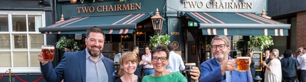 An image showing 4 people stood outside at The Two Chairmen behind a red ribbon holding drinks up in the air in celebration.