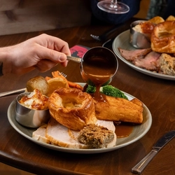 A table setting inside a cozy restaurant or pub, where two people are seated and sharing a meal. The table holds plates of food, including what appears to be a roast dinner, along with wine glasses, cutlery, and condiments. One person reaches toward the food while the other sits opposite. .