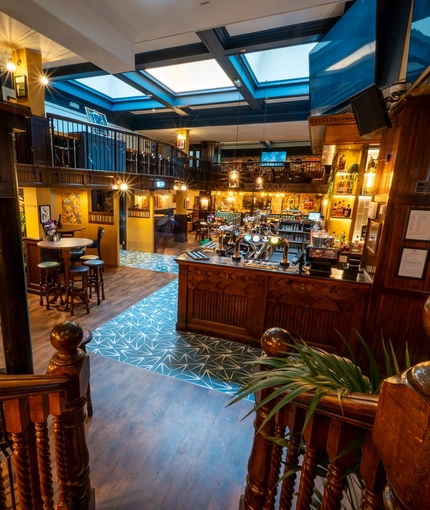 The central bar area inside the Old School House, with TVs above the bar, large sky lights in the ceiling, and restaurant seating areas on the lower and upper floors, with a balcony overlooking the bar.