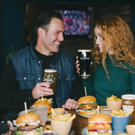 A lifestyle image of 4 friends sat within the interior restaurant seating area enjoying Burger dishes and various drinks available at Urban Core venues.