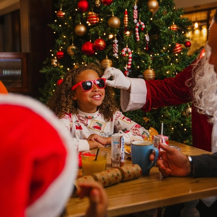 A child wearing Christmas-print pyjamas and red novelty sunglasses sits at a festive breakfast table in front of a decorated Christmas tree. A person dressed as Santa Claus adjusts the sunglasses while other diners sit around the table. The table contains breakfast plates, a mug, a glass of orange juice, juice cartons and Christmas crackers. The tree is decorated with red and gold baubles, candy cane ornaments and warm fairy lights, creating a festive restaurant setting.