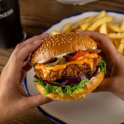 Two hands holding a Spicy Franks Mayo Chicken Burger over a plate on a wooden table.
