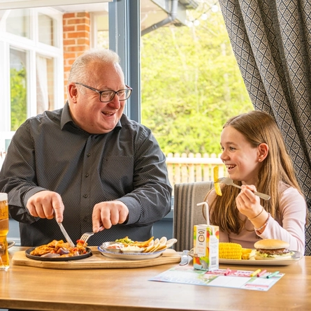 Three people comprised of a man, woman and a child are seated at an indoor table with plates a food. The child is holding up a fork with a chip on it.  They are smiling and enjoying conversation.