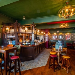 The interior restaurant seating area and wood panelled bar at the Two Chairmen, with upholstered chairs and bar stool seating, wooden flooring, and framed artwork on the floral wallpapered walls.