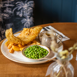 An advertising image of a plated Cod & Chips dish sat on a table within the interior restaurant seating area at a Chef & Brewer venue.