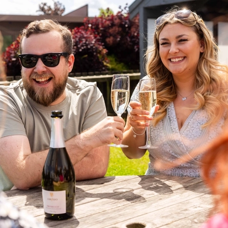 Four people sitting at a wooden table in a sunny beer garden. Two people are holding glasses of prosecco, and the bottle sits on the table in front of them.