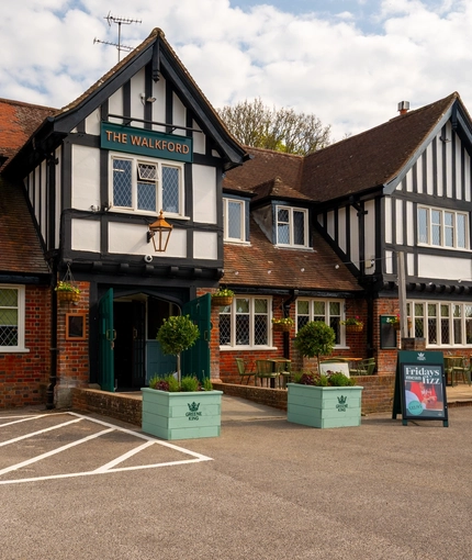 The exterior facade, signage, and seating area at The Walkford, with flower baskets hanging on the walls, string lights above the tables and chairs, and accessible parking spaces near the entrance.