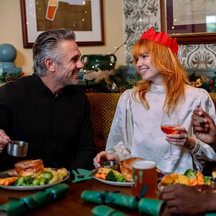 A person wearing a red paper crown sits at a table in a festively decorated pub, holding a glass of rosé wine beside a plate of food containing prawns, potatoes and salad leaves. Additional roast dinners, glasses of red wine and green Christmas crackers are arranged on the table. A Christmas wreath, seasonal garlands, fairy lights, patterned wallpaper and exposed brickwork create a festive dining setting.