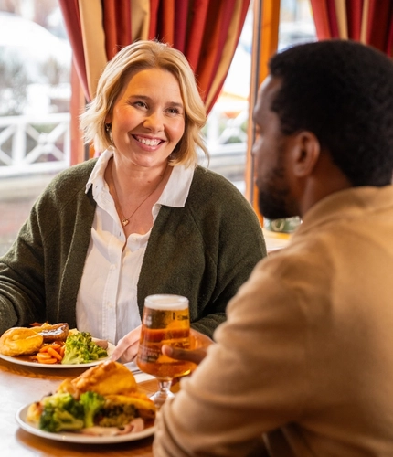 Two people sitting at a restaurant table with plates of roast dinner in front of them. One person is holding a glass of wine, the other is holding a glass of beer, and menus sit on the table along with salt and pepper.