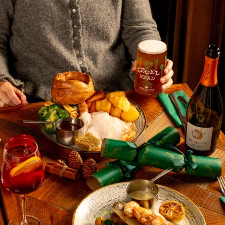 A person is seated at a wooden table. The table is decorated with festive decoration and is  holding a plated turkey dinner.  Only the torso and arm is visible in the image. The person is holding a pint of Level Head.
