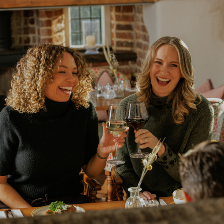 An image focusing on 2 customers celebrating with a cheers sat with friends enjoying starter dishes from the Prix Fixe menu sat at a table within the interior restaurant seating area of The Crown.