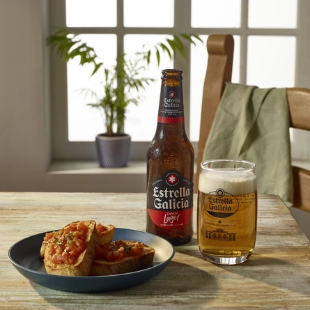 A bottle and half a pint of Estrella Galicia with a ciabatta and tomato plated dish sat on a wooden table.