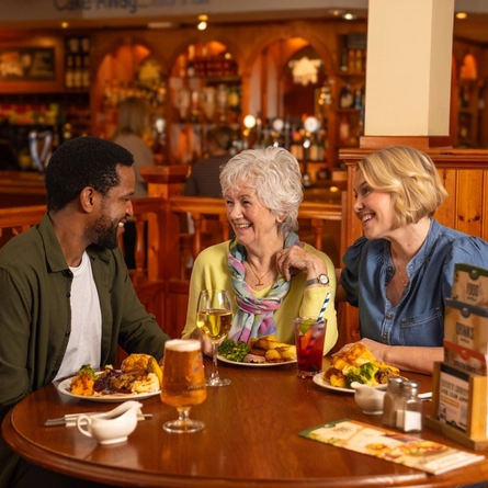 Three people sitting at a wooden restaurant table with plates of roast dinner in front of them. A glass of wine, a glass of beer, and a glass of fizzy drink also sit on the table, along with menus, salt, pepper, and jugs of gravy.
