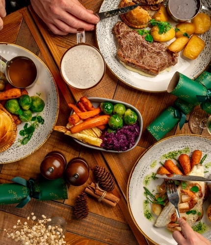 A overhead view of a festive decorated wooden table holding a variety of main meals and drinks.  There are hands holding cutlery as if to take a piece.
