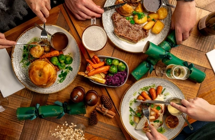 A overhead view of a festive decorated wooden table holding a variety of main meals and drinks.  There are hands holding cutlery as if to take a piece.