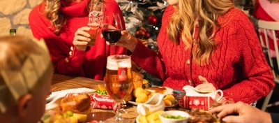 Two people in festive red jumpers sit at a Christmas dining table in a pub decorated with a snow-covered Christmas tree and red and gold baubles. One wears a reindeer antler headband while the other wears a paper crown from a Christmas cracker. They raise a pint of lager and a glass of red wine in a toast, with a traditional Christmas dinner, drinks and crackers visible on the table.