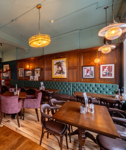 An interior restaurant seating area at The Plough, with upholstered chairs and booth seating, and framed artwork on the wood panelled wall.