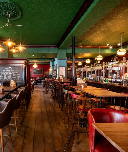 The bar and interior restaurant seating area at the King's Head, with dark wood floors; green, textured wallpaper on the ceiling; and a long, red, upholstered booth seat along one wall by the window.