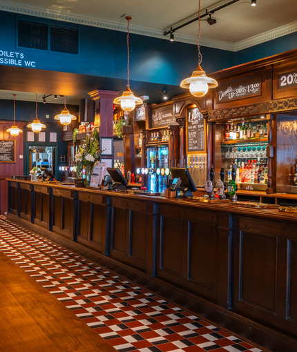 A close up view of the wood panelled bar at the Scholars Arms in Southampton; with patterned tile flooring in front of the counter, and a TV on the wall.