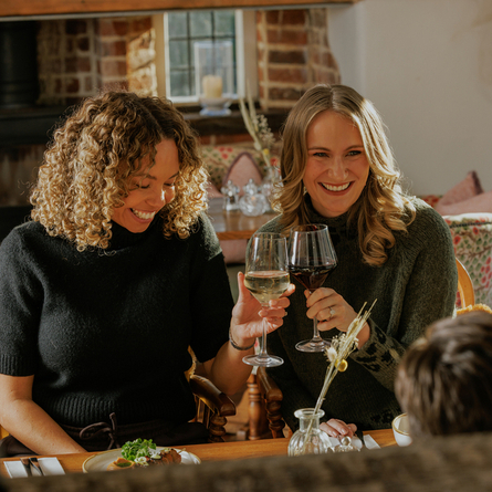An image focusing on 2 customers celebrating with a cheers sat with friends enjoying starter dishes from the Prix Fixe menu sat at a table within the interior restaurant seating area of The Crown.
