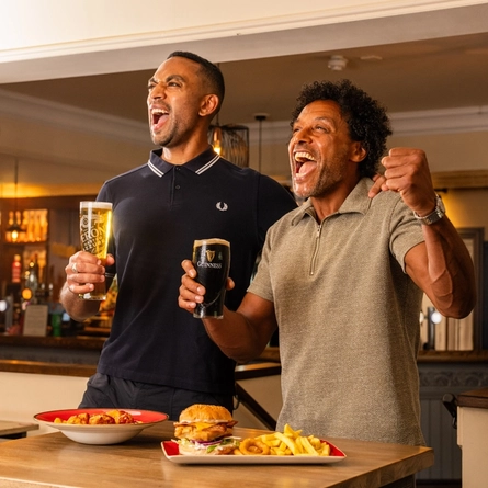 Two people standing beside a high table in a pub, holding a pint of lager and a pint of Guinness. Plates of food, including a burger with chips and a pasta dish, are on the table. The pub interior features a stocked bar, exposed brickwork and warm lighting in the background.