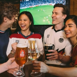 An image showing a group of friends stood at a table watching a football match and enjoying a selection of drinks while celebrating within the interior seating area at a Greene King venue.