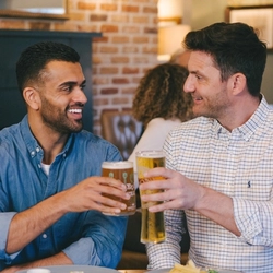Two people sitting at a restaurant table, clinking together their glasses of beer. Two plated meals, cutlery, and salt and pepper mills sit on the table.
