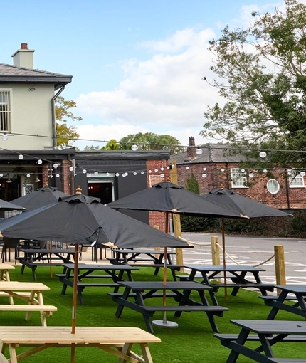 The exterior facade, signage, and beer garden seating area at The Carousel, with wooden picnic table, shade umbrellas, and string lights.