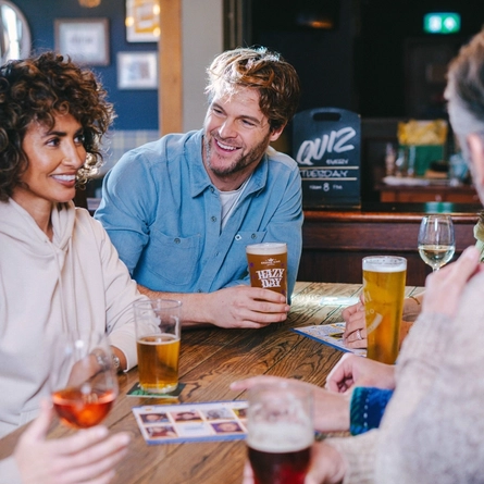 A lifestyle image of a group of friends sat at a table enjoying drinks during a Quiz event at a Greene King Pub.