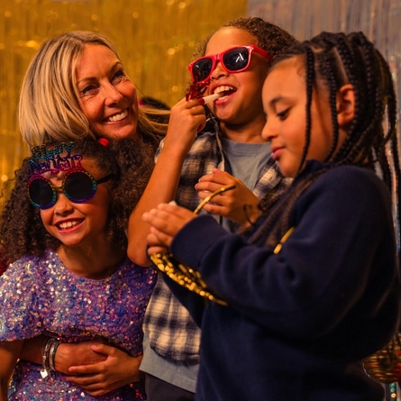 A festive New Year’s celebration taking place in front of silver and gold metallic party backdrops. Several people wear novelty party glasses and hold red party blowers, while one person in the centre wears oversized colourful sunglasses and a sequinned outfit. A decorated Christmas tree with warm fairy lights is partially visible in the foreground, and bright decorative lighting creates a vibrant party atmosphere.