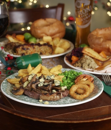 An advertising image focusing on a Christmas Day Stilton Steak Carvery dish with various plated Festive Main dishes in the background and a selection of drinks available at a Pub & Carvery venue.