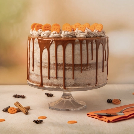 The Pumpkin Spiced Latte cake, served on a glass cake stand which sits on a table strewn with cinnamon sticks, star anise, and mini pumpkin decorations. A napkin and cake slicer sit on the table next to the cake stand.