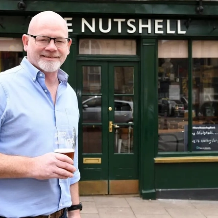 A person standing in front of a pub while holding a pint of lager.