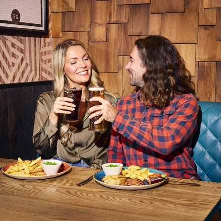 An image showing 2 friends sat at a table with drinks and celebrating with a cheers while enjoying food dishes within the interior restaurant seating area at a Flaming Grill venue.