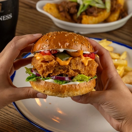 Two hands holding a Spicy Franks Mayo Halloumi Burger over a plate on a wooden table.