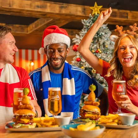 Three football fans sit around a wooden table in a sports pub decorated for Christmas, celebrating while watching live football on large screens. A Santa hat, reindeer antlers and team scarves add to the festive atmosphere, with a frosted Christmas tree in the background. Burgers topped with onion rings and pickle skewers, chips and glasses of beer are served on the table, creating a lively Boxing Day matchday dining scene.